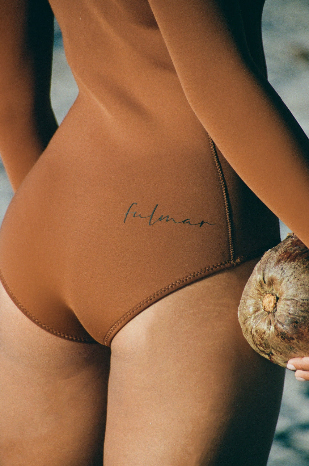 Brown swimsuit with 'Fulmar' branding held by a person on a beach.